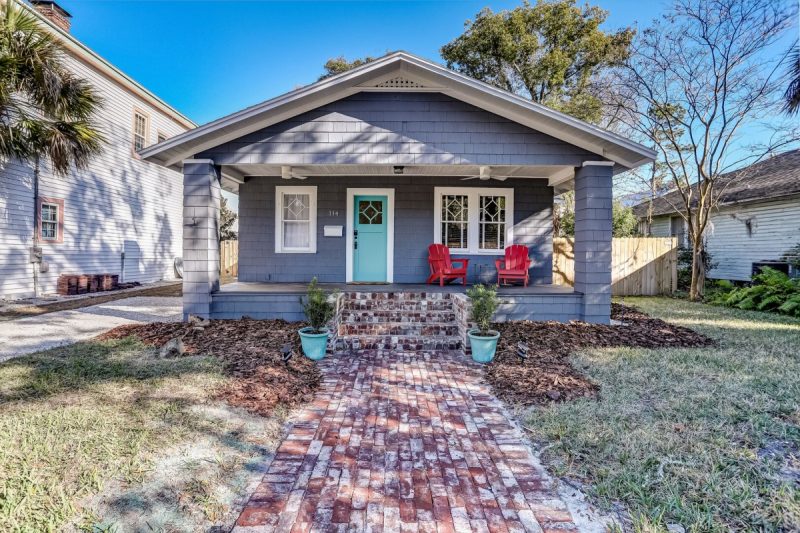 Grey cottage home with turquoise accent door and red Adirondack chairs sitting on a spacious front porch. Featuring a beautiful brick walkway and steps 