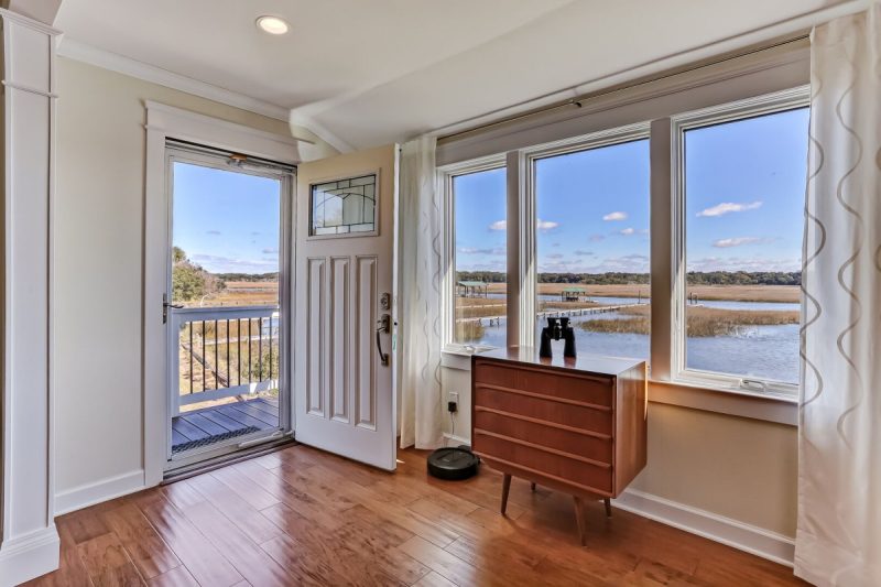 Front entryway in living room showing large bay windows with a marsh view. Living room is bright with hardwood floors, mid-century-modern furniture piece, and Roomba vacuum